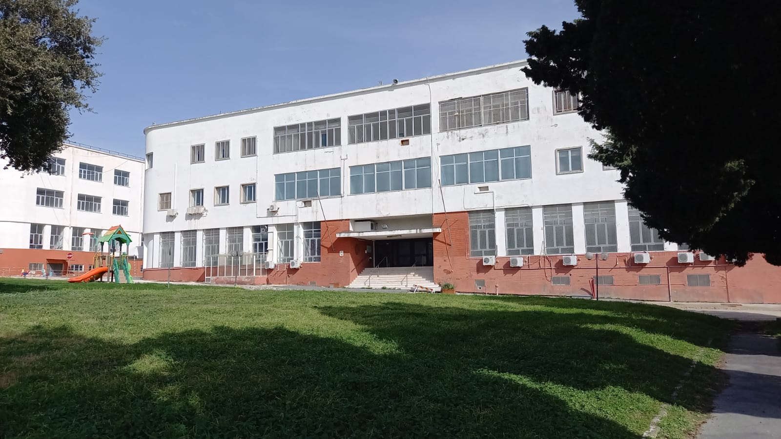 A long white three-storey rationalist dormitory building with ribbon windows and a red brick base, seen across grass, with a children’s playground at the left.