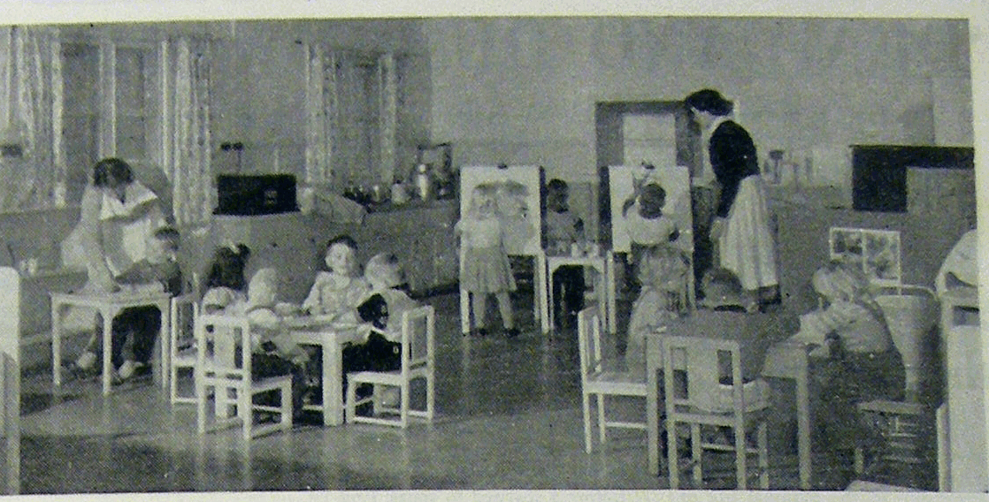 Children and adults in a kindergarten room with small tables and easels.