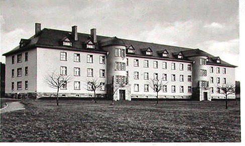 A long four-storey barrack block with dormer windows, seen across a lawn with bare young trees.