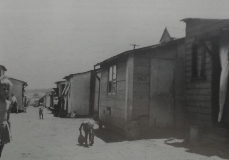Rows of railway camp huts beside an unpaved lane, with people outside.