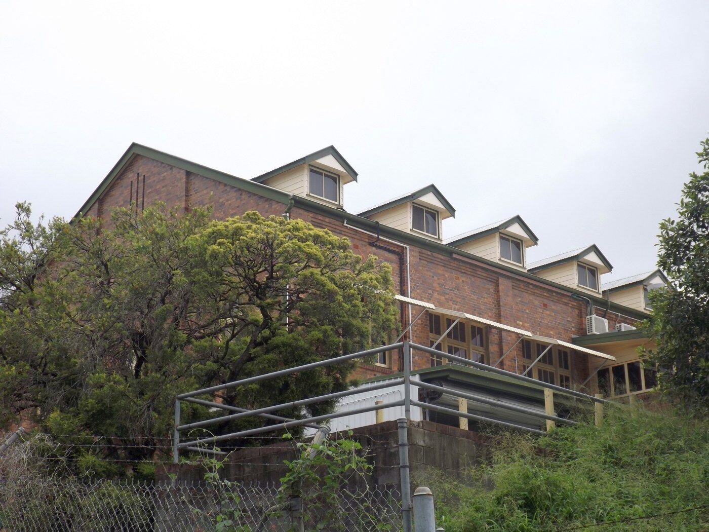 Two-storey red brick factory building with dormer windows on a slope behind a wire fence.