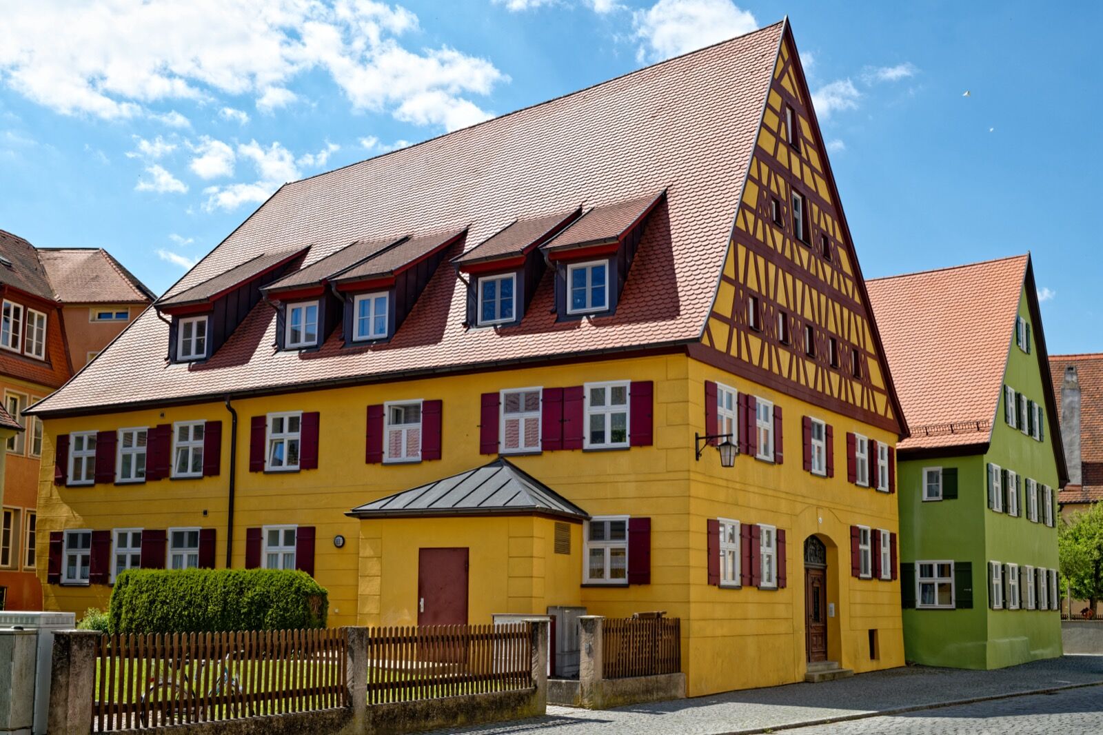 A tall yellow-rendered house with a steep tiled roof, four dormer windows, red-brown shutters and a timber-framed gable, standing on a cobbled street corner.