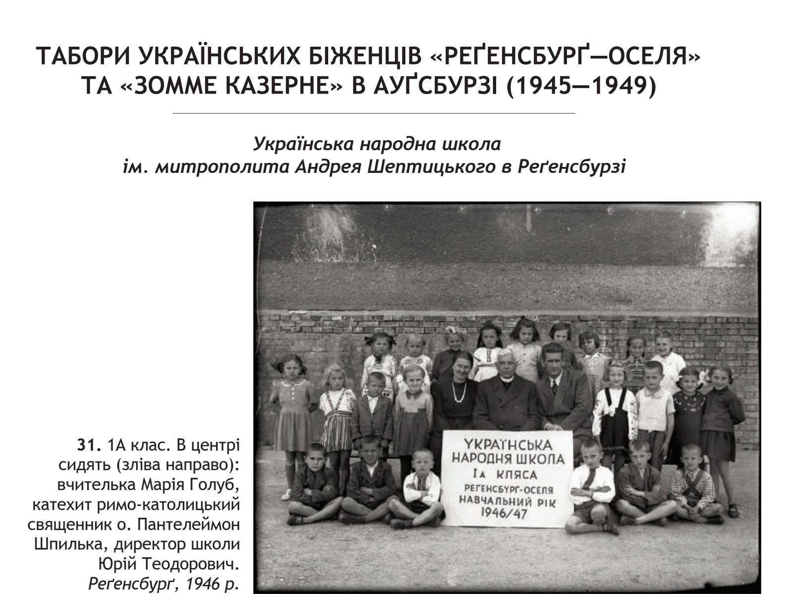 A class of about thirty children standing and sitting in rows before a brick wall, with three adults seated in the centre and a hand-lettered placard propped in front of them.