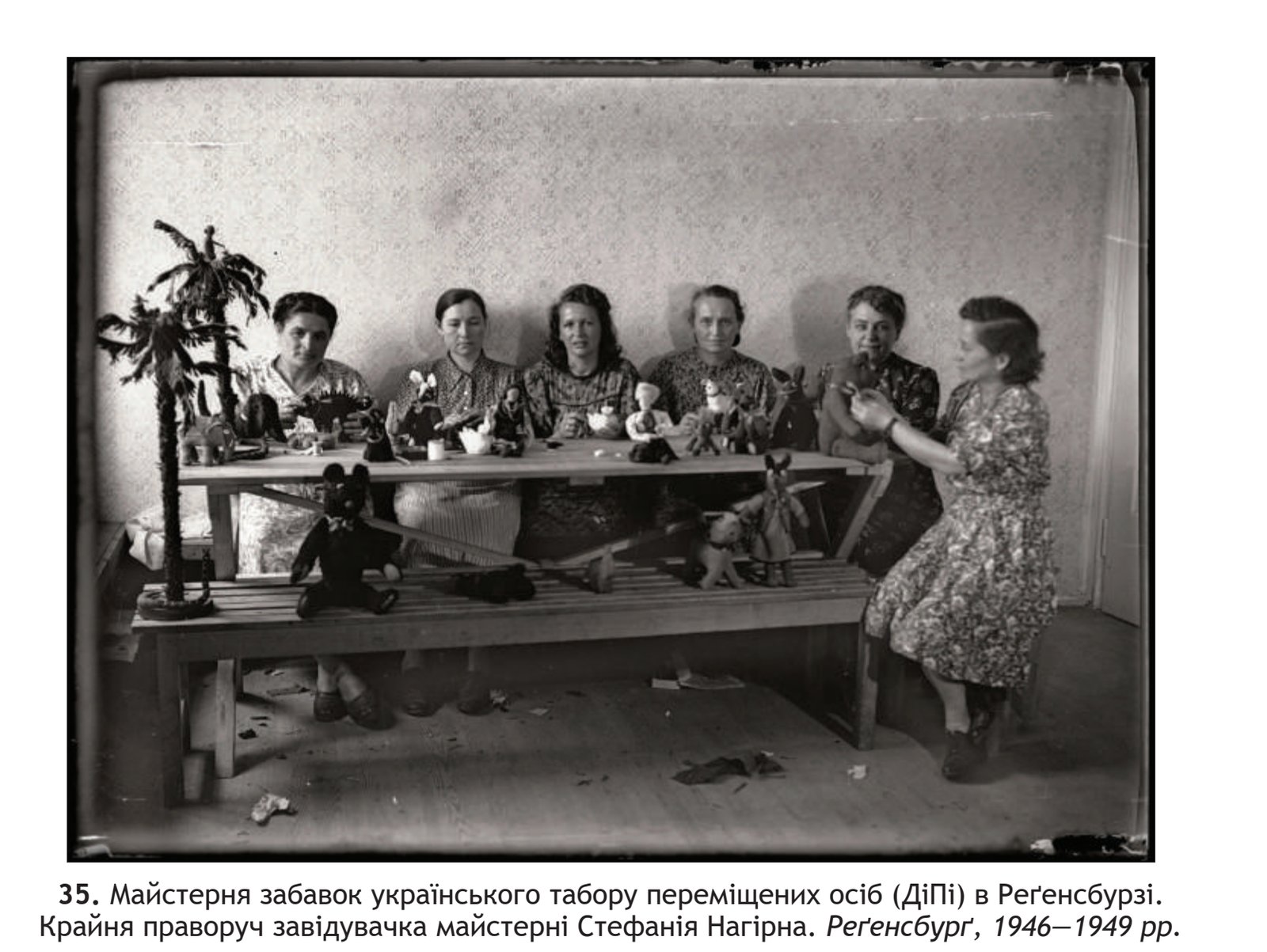 Six women seated behind a long wooden table covered with small stuffed toys and dolls, with more toys and a model palm tree on the bench in front.