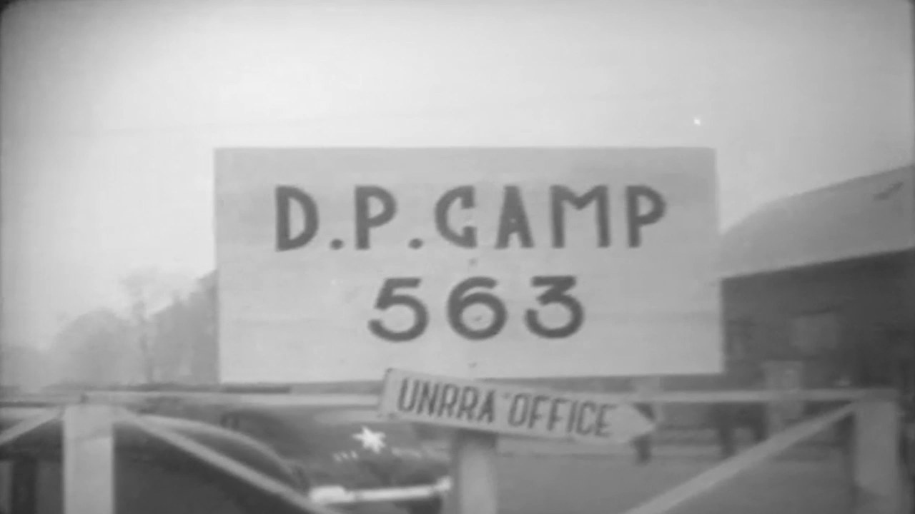 A painted board reading D.P. CAMP 563 above an entrance barrier, with a smaller hanging sign reading UNRRA OFFICE.