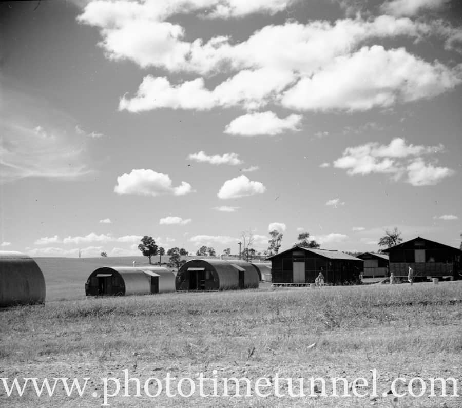 Nissen huts and gabled timber huts standing in a paddock under a cloudy sky, with a few figures beside them.