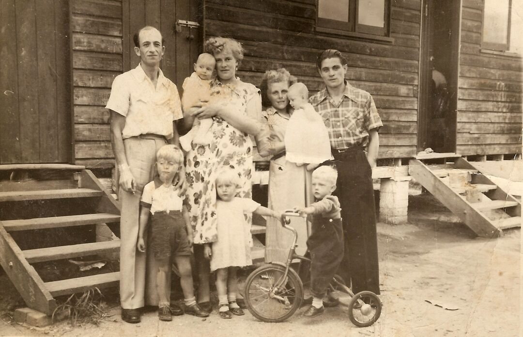 Four adults and five children outside a timber hut; tricycle in foreground.