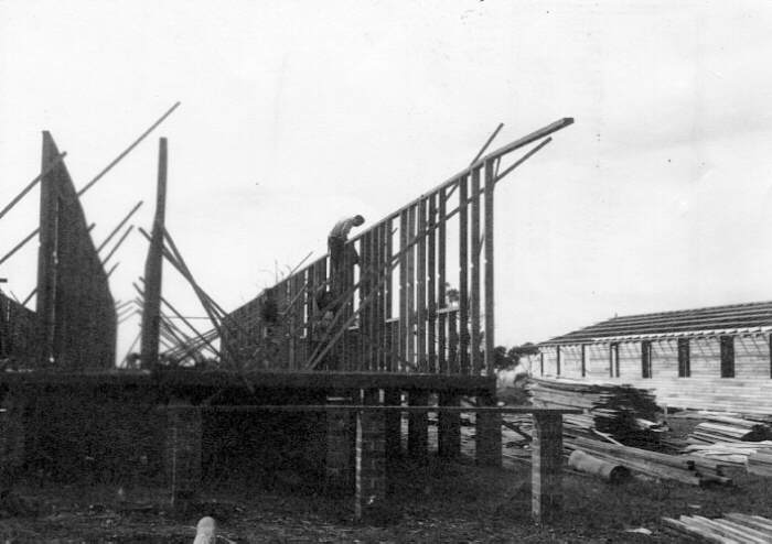 Workers erect timber framing alongside long hut.