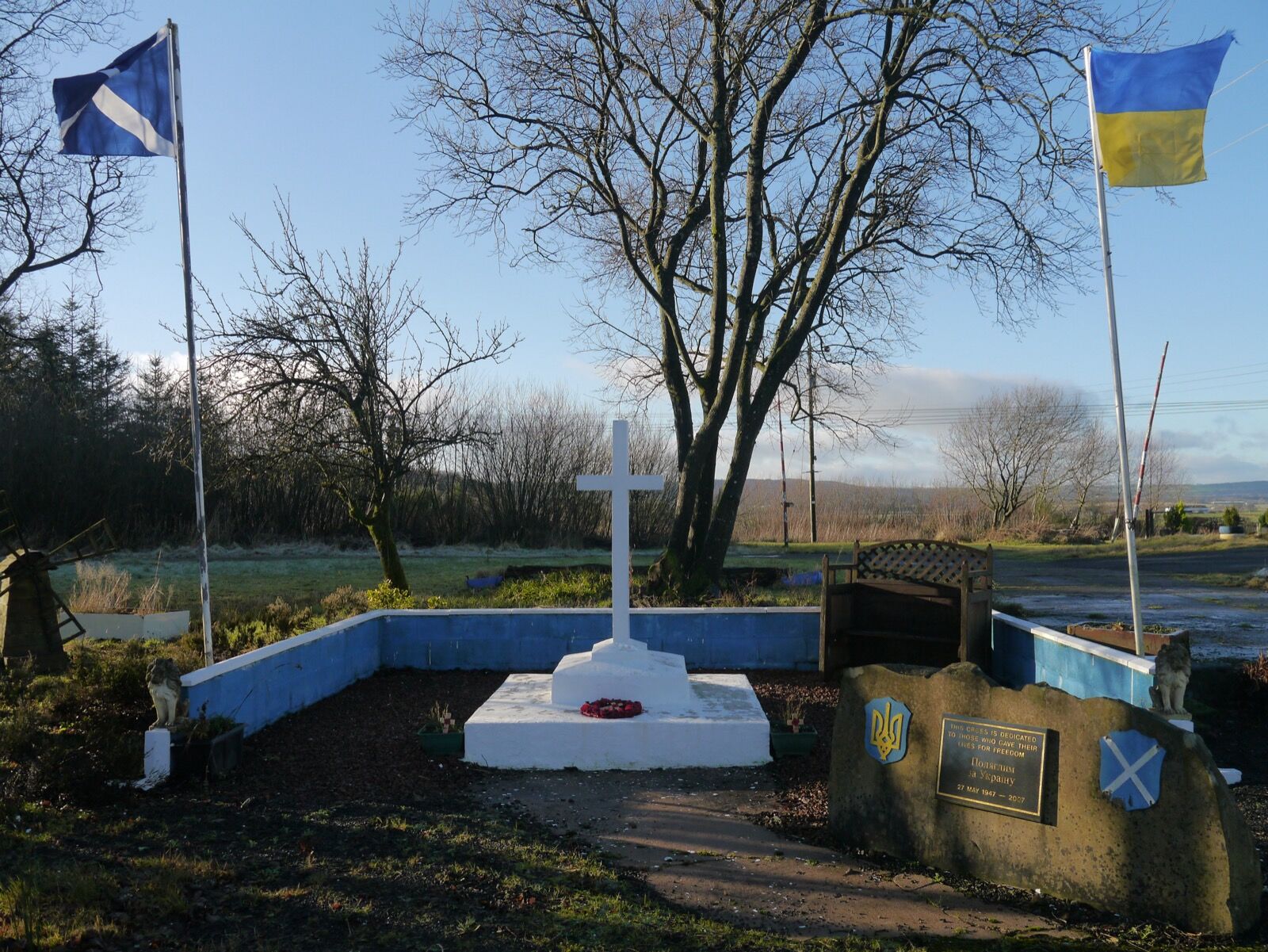 A white cross in a low blue-painted enclosure at Hallmuir, between the Scottish and Ukrainian flags, with an inscribed plaque on a stone beside it.