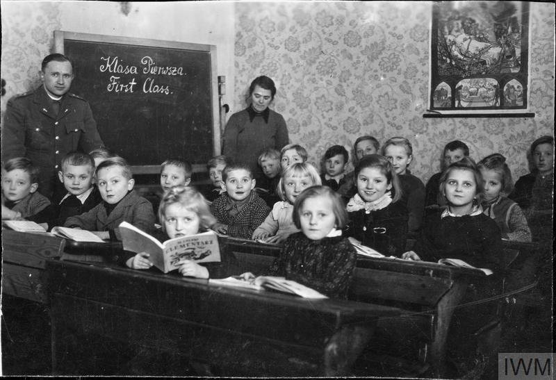 About twenty children seated at wooden desks in a papered room, with a blackboard behind them chalked Klasa Pierwsza and First Class, a man in uniform on the left and a woman standing behind the desks.