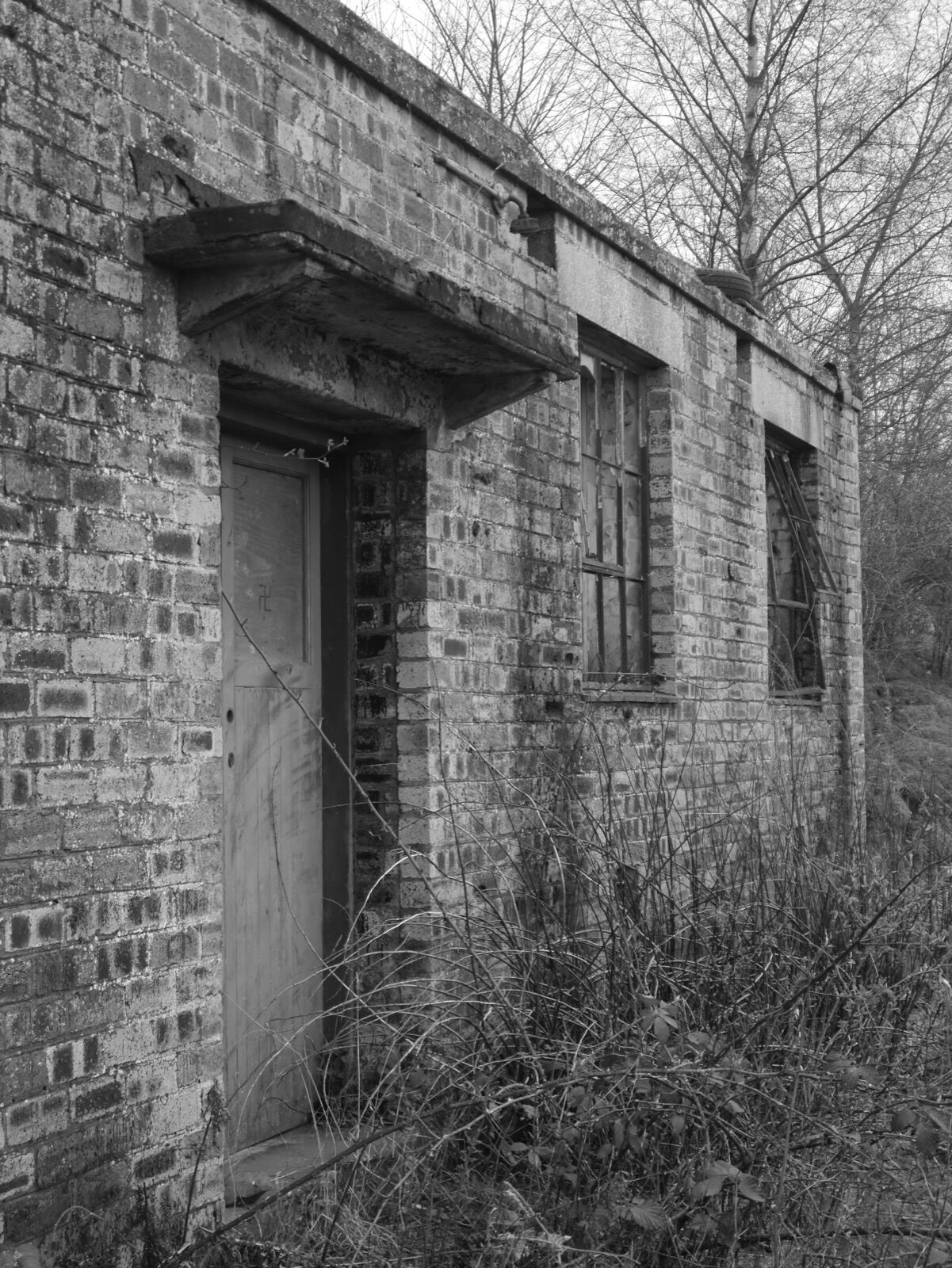 Black and white photograph of a derelict single-storey brick building with a concrete canopy over its doorway and broken metal-framed windows, overgrown with scrub.