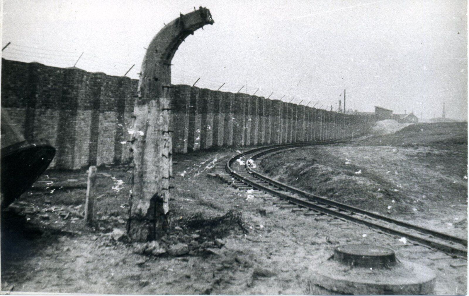 A long concrete wall with barbed wire along the top, a tall curved lamp post in front of it and a narrow-gauge railway track curving away over bare ground.
