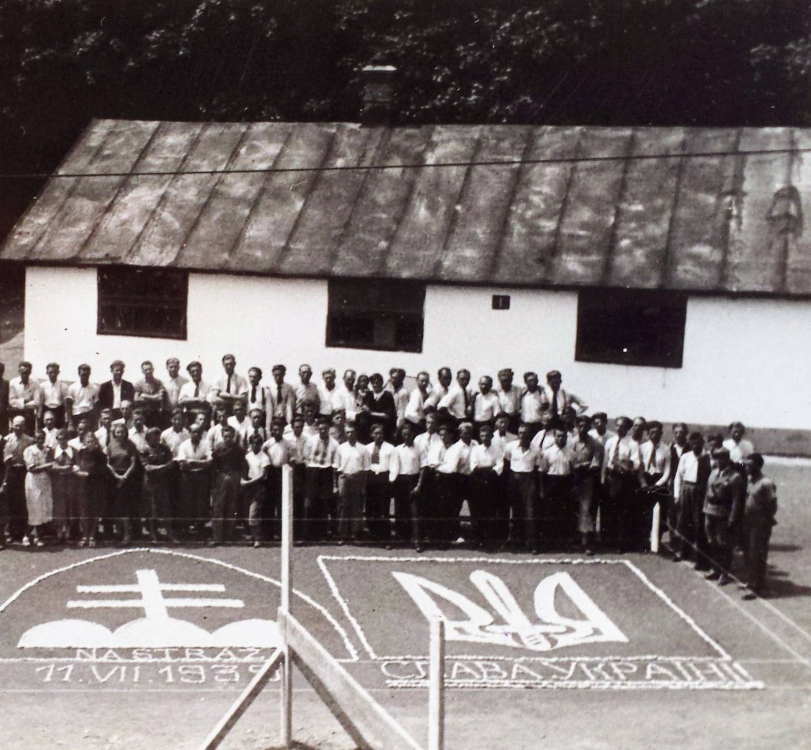 Men of the camp drawn up in front of a long white single-storey barrack, with a Slovak double cross and a Ukrainian trident laid out in white stones on the ground in front of them.