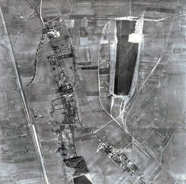 Black-and-white vertical aerial photograph of flat farmland with a village street, a long dark flooded gravel pit and rows of small buildings, the name Karlsfeld printed across it.