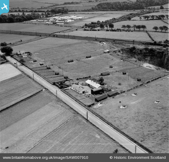 Oblique aerial view of farmland and hatchery, with a regular hutted compound beyond a railway.