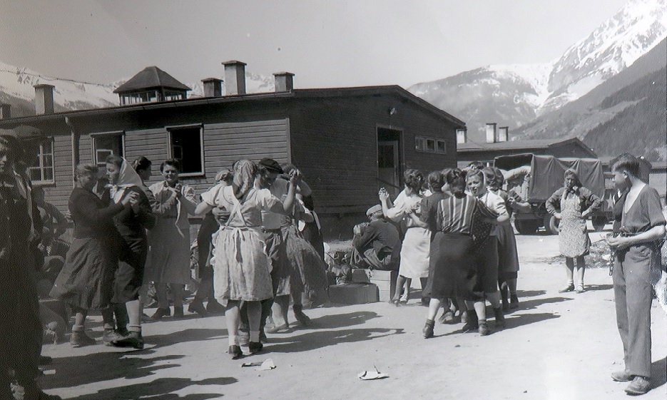 People dancing in pairs on open ground in front of a long timber barrack at Landeck, with mountains and an American army lorry behind.