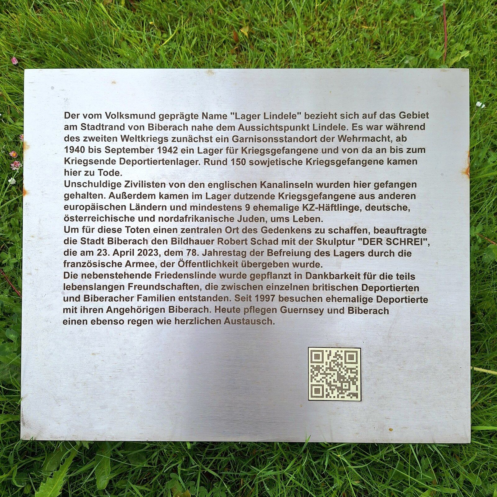 Steel plaque lying on grass with German text explaining the history of Lager Lindele and the sculpture Der Schrei.