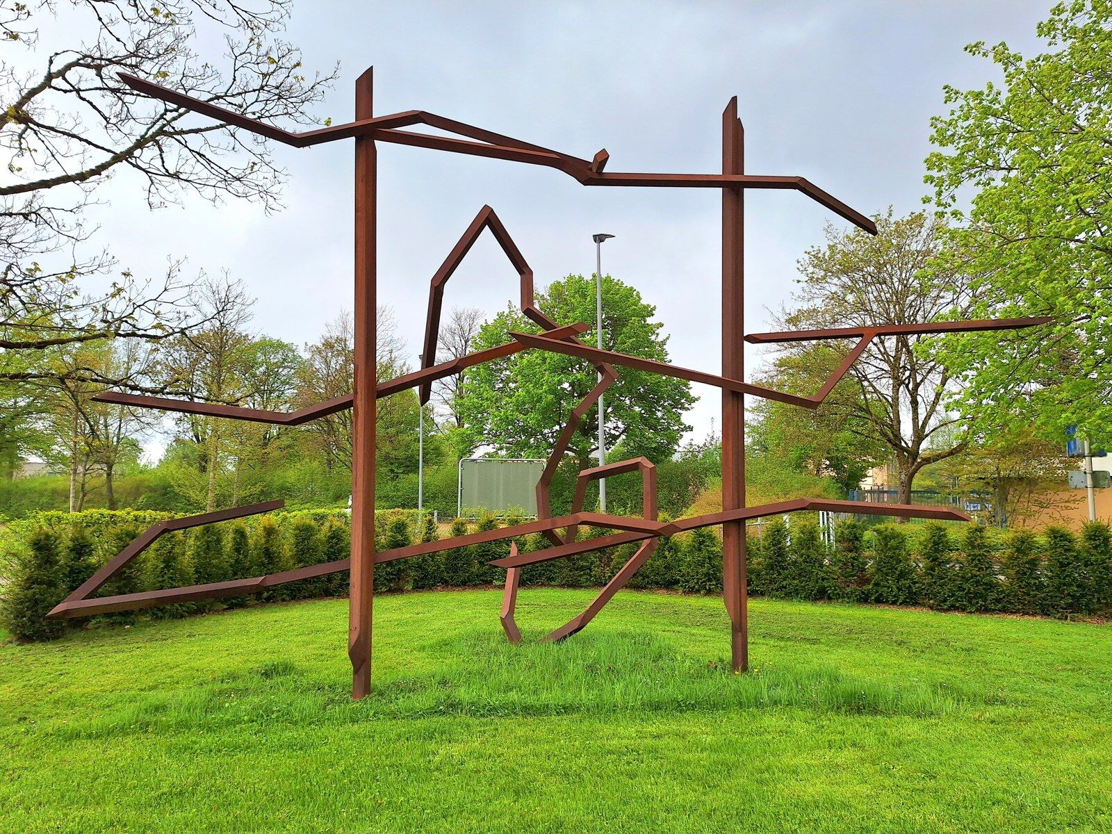 Large rust-coloured steel sculpture of angular lines standing on a lawn in front of a hedge and trees, outside the former Lager Lindele in Biberach.