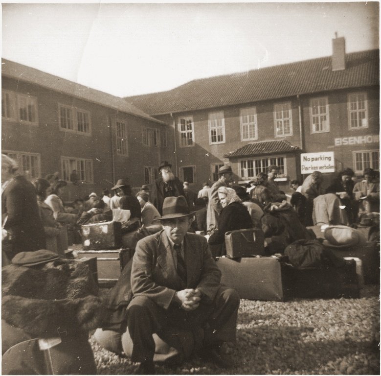A crowd of men, women and children sitting on suitcases and bundles in a yard in front of two-storey brick buildings, with a sign reading No parking on the wall behind.
