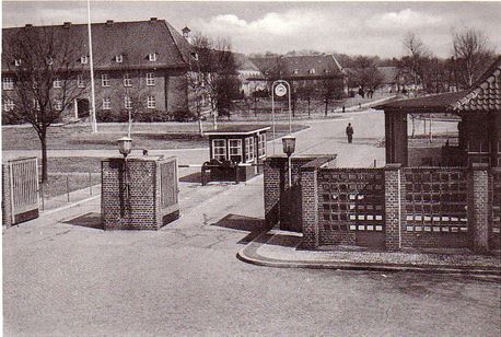 The entrance to Camp Grohn: brick gate pillars, a guard hut and a barrack block behind, photographed in black and white.