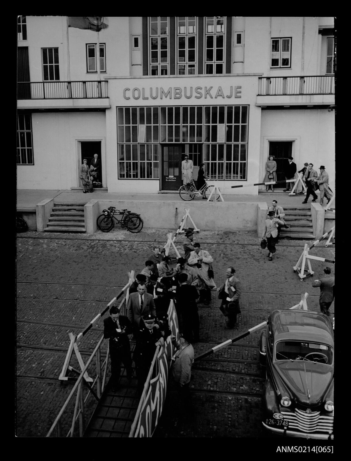 Emigrants carrying bags walk up a gangway past barriers in front of a white building lettered Columbuskaje.