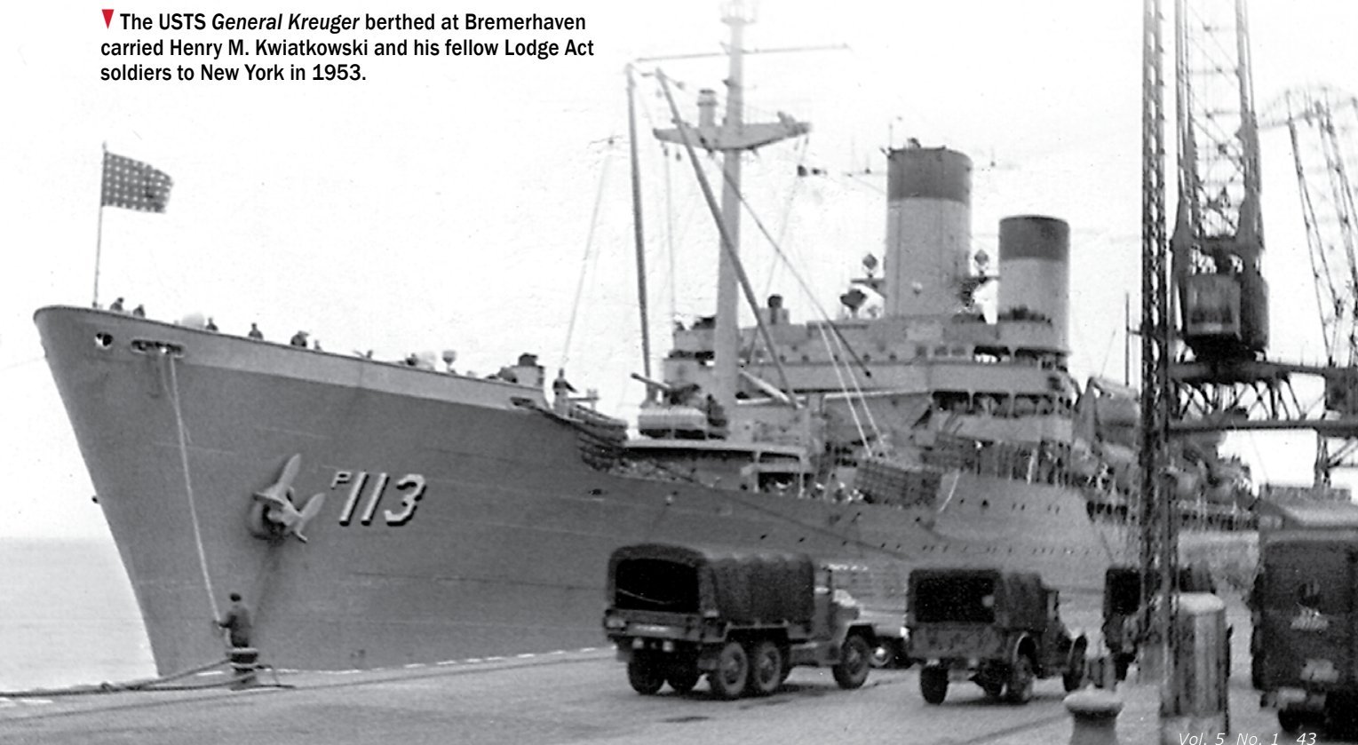 A large grey troopship berthed at a quay, her bow marked with a hull number, with army lorries and cranes on the dockside.
