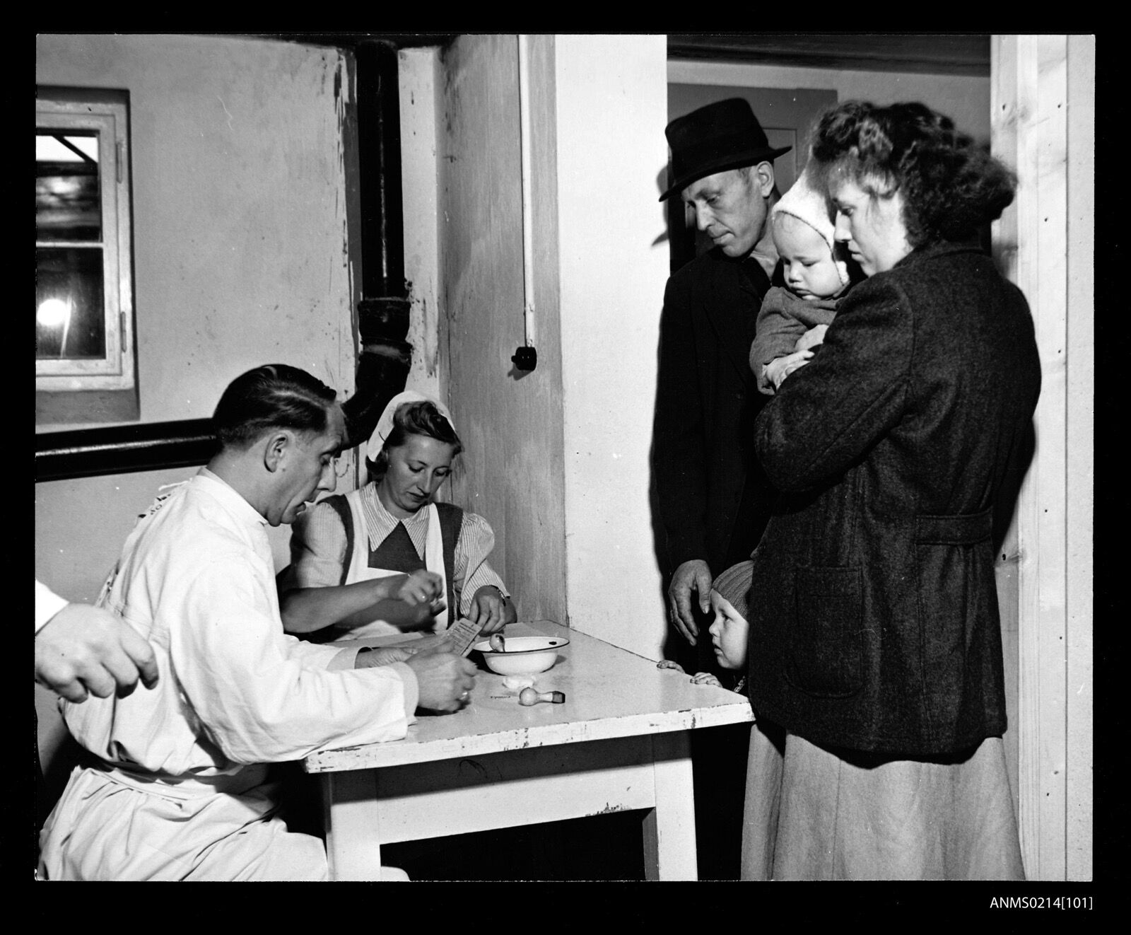 A family with a child in arms standing before a doctor at a table during a medical examination in a camp room at Lesum.