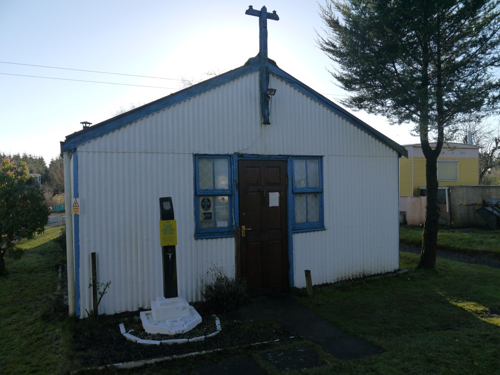 A long low hut of corrugated iron and brick under a pitched roof, with a small bell and a cross above the entrance, standing in open ground.