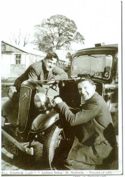 Two men stand beside a vehicle with a low camp building behind them.
