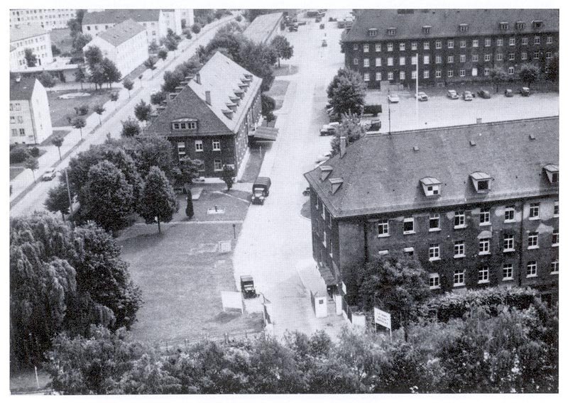 A black-and-white aerial view of barrack blocks with pitched roofs along a tree-lined street, with parked cars, a lorry and a gate with sentry boxes.
