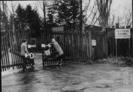 Two people at a wooden paling gate beside a sign reading Lettische Kolonie, with barracks and bare trees behind.