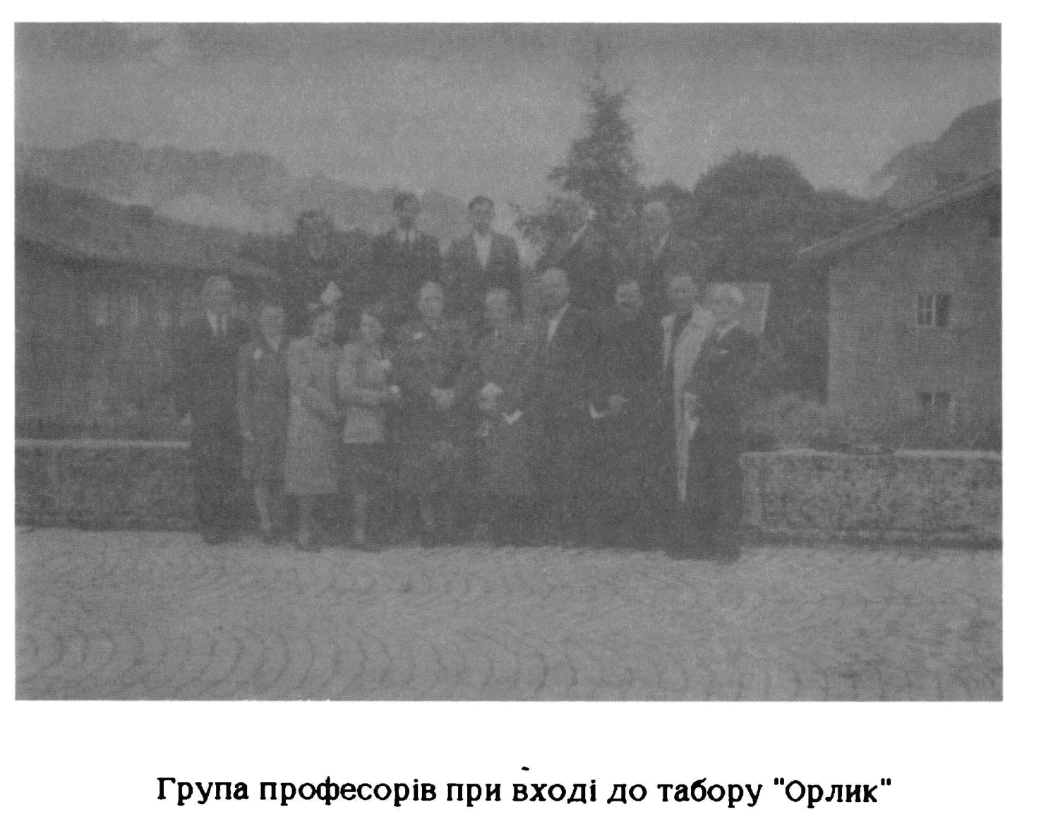 A group of about eighteen men and women in coats standing on a cobbled forecourt between two camp buildings, with wooded mountains behind.