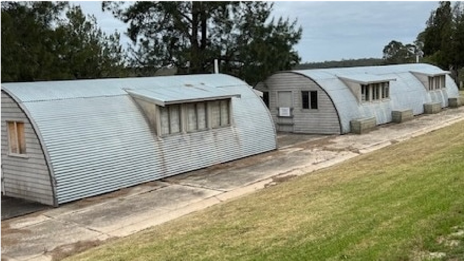 Three curved corrugated-iron huts in a row on a grassed slope, each with a weatherboard end wall, a door and a hooded window, with gum trees behind.