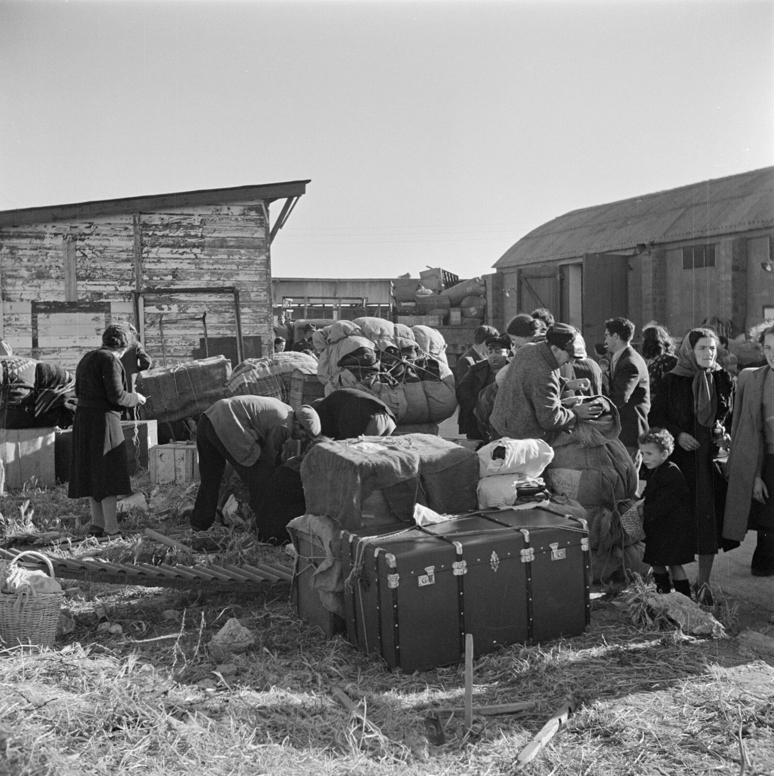 Newly arrived immigrants standing among suitcases and bundles outside low huts at the St Luke’s transit camp near Haifa.