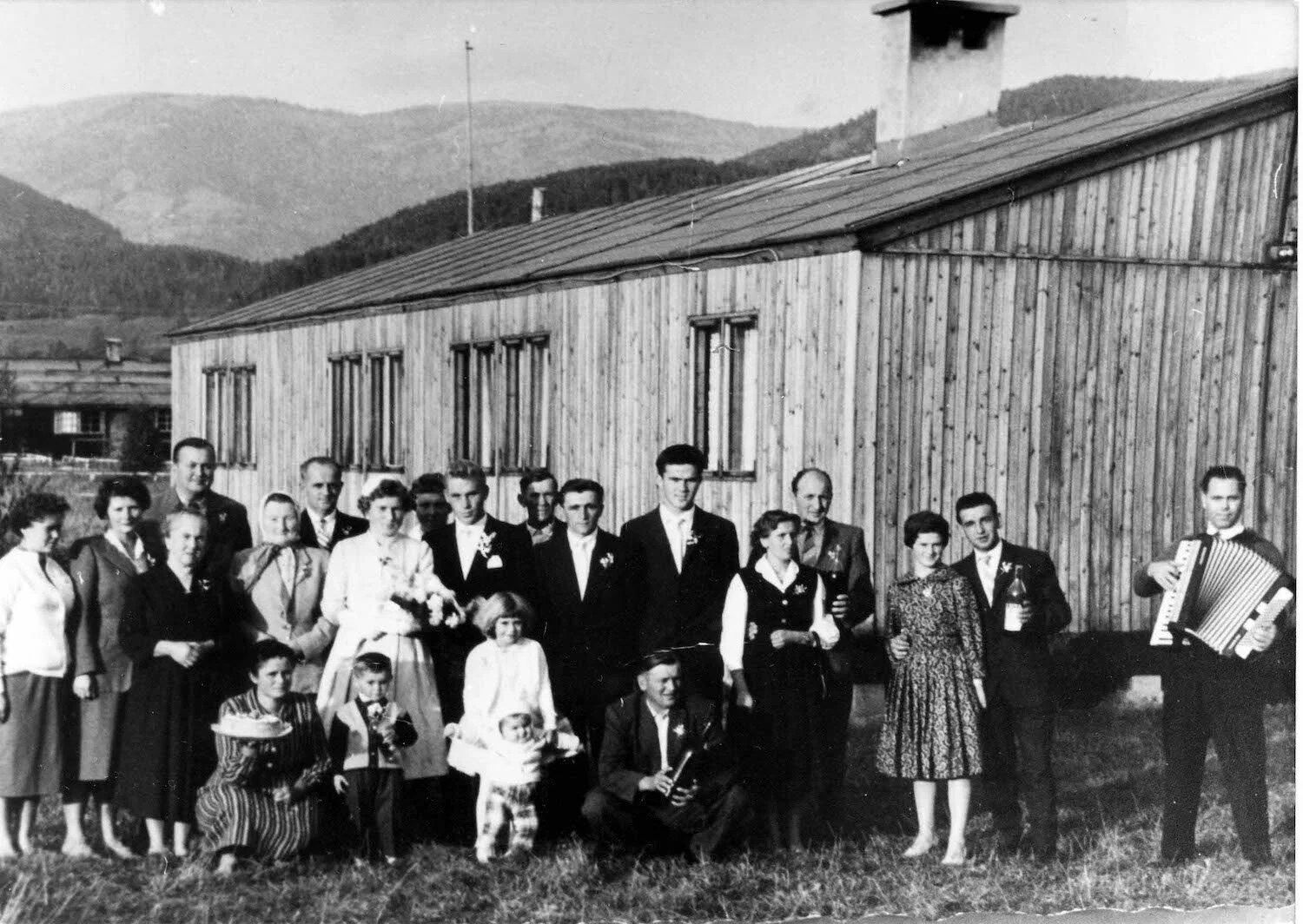 A wedding party of about twenty adults and children standing before a timber hut, one man holding an accordion.