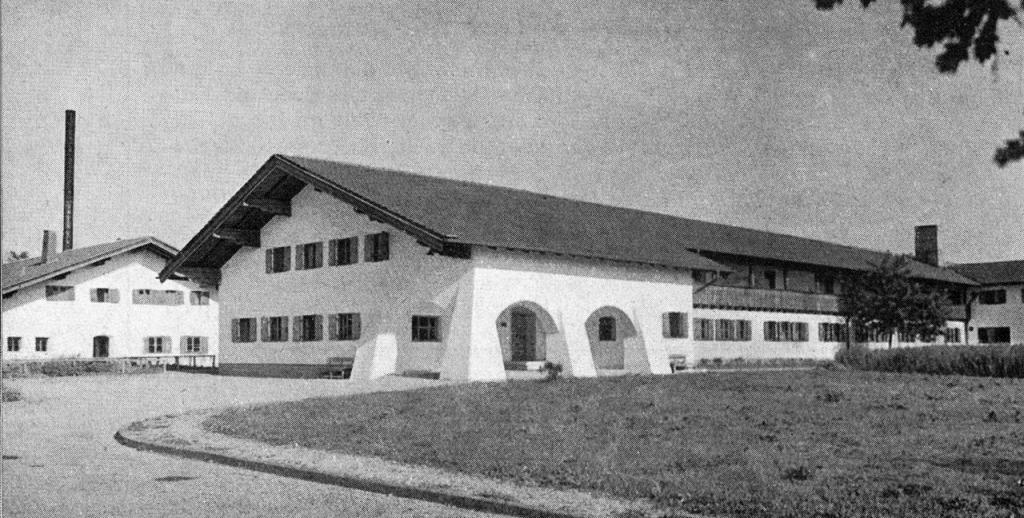 Black-and-white photograph of a long white barracks building with a broad overhanging roof and an arched entrance, standing on open grass.