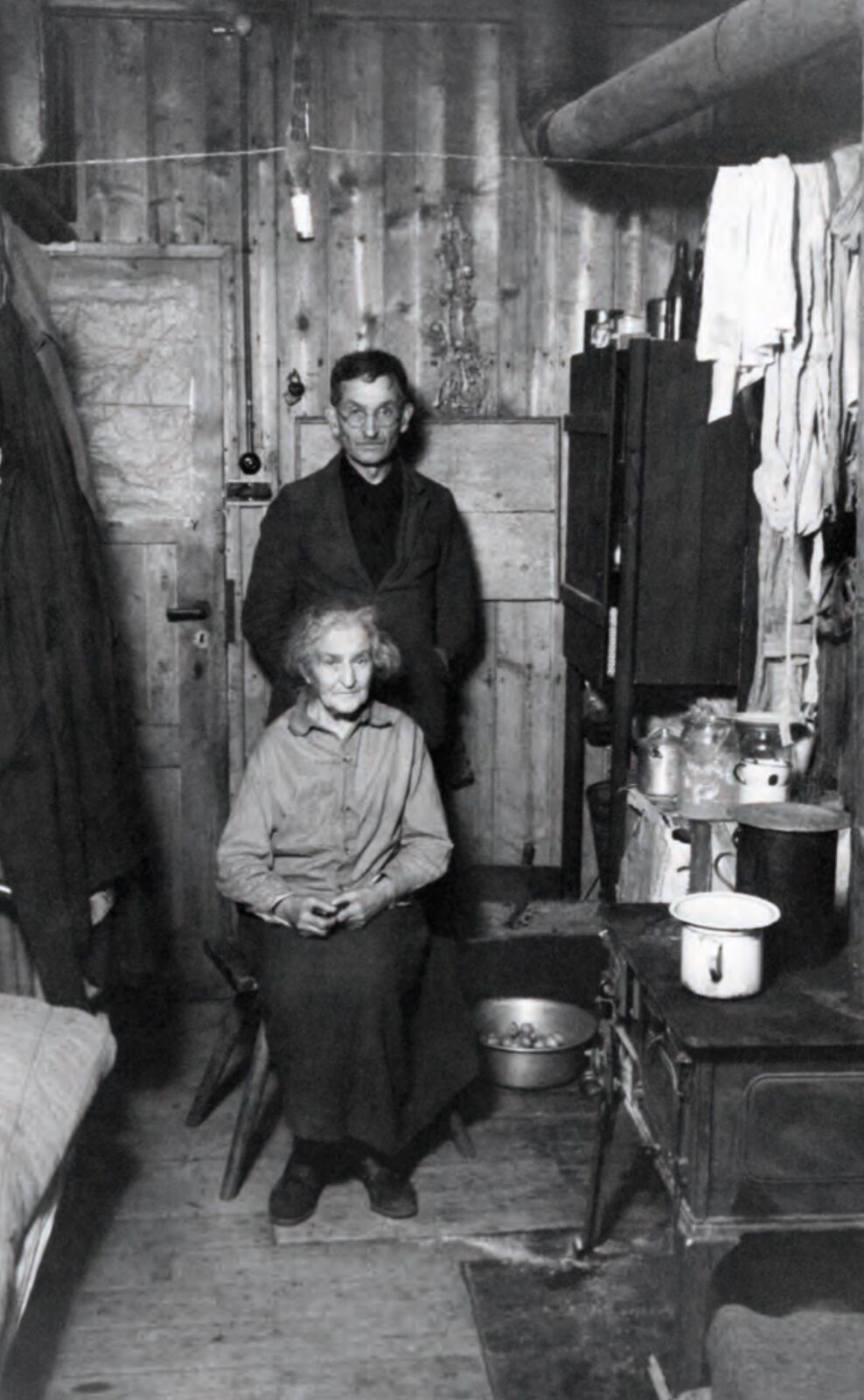 Black-and-white photograph of 1946 showing an elderly couple in a wooden barrack room with an iron stove, pans and clothing hung on the boarded wall.