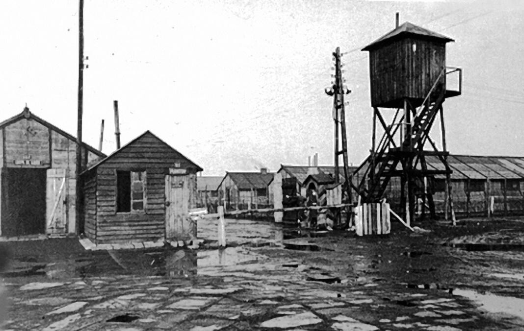 Wooden barracks, a sentry box and a raised timber watchtower standing over waterlogged ground at the Waterschei camp.