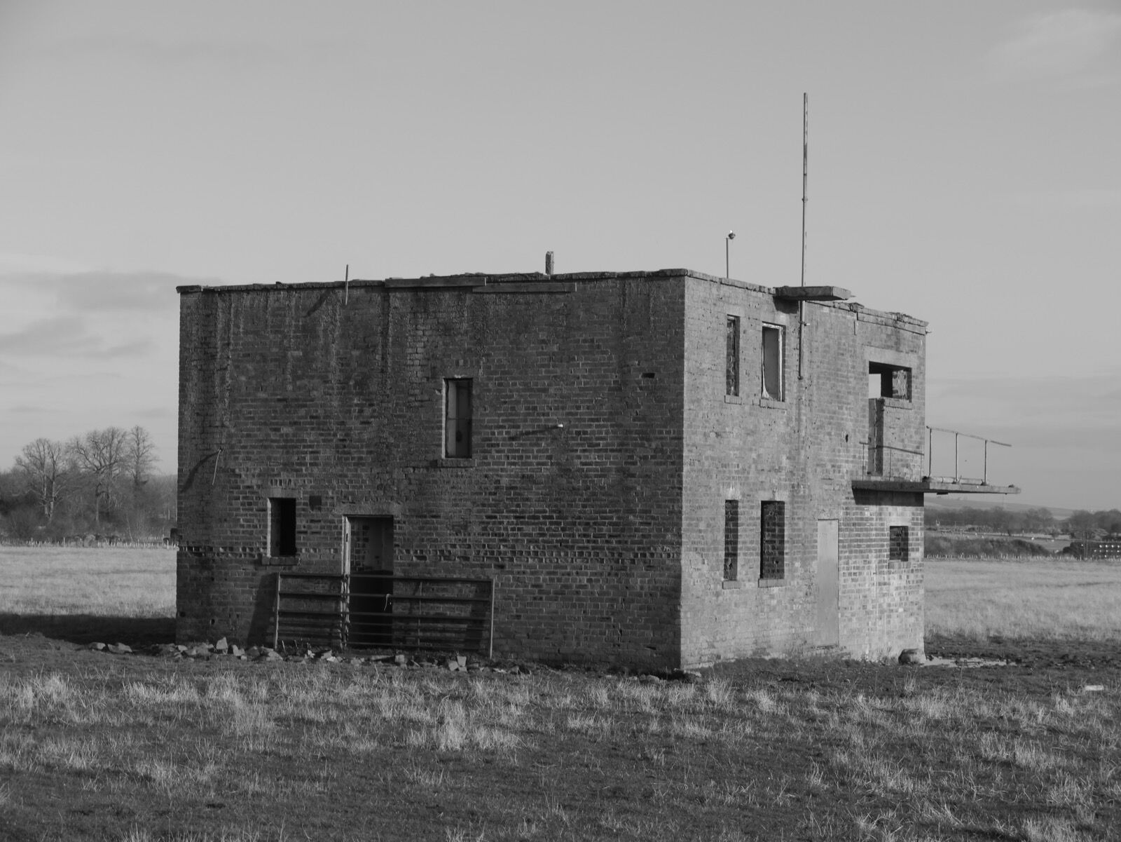 Black and white photograph of a two-storey brick watch office standing alone in a grass field, its windows empty and a mast on the roof.