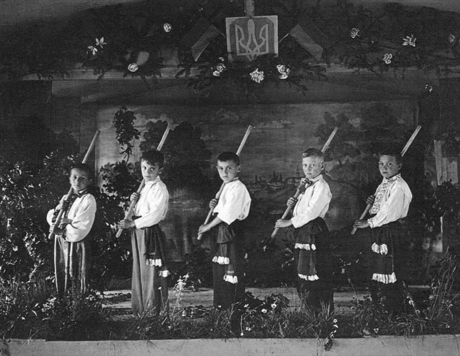 Five boys in embroidered shirts holding scythes stand on a stage in front of a painted rural backdrop with a trident emblem above.