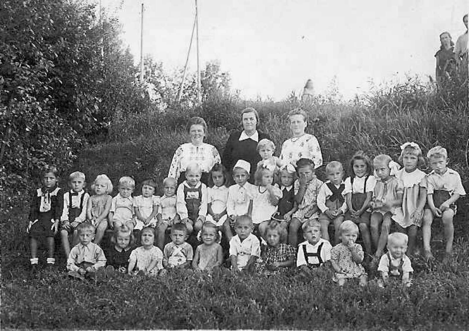 Black-and-white group photograph of about forty small children with three women on a grassy bank.
